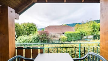 Ein Holzbalkon mit Tisch und Stühlen, mit Blick auf ein Haus mit rotem Dach und üppiger Bepflanzung.