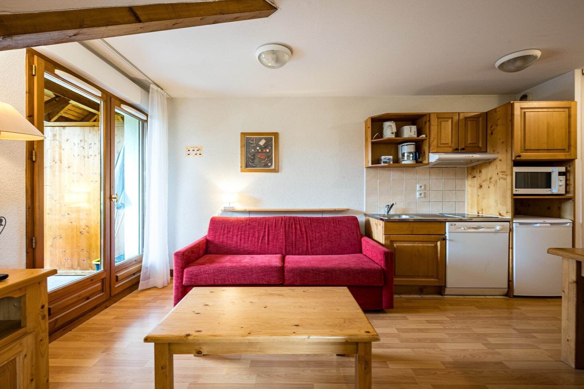 Eine kompakte Küche mit Holzausstattung, einer Mikrowelle und einem Geschirrspüler. Im Wohnbereich befinden sich ein rotes Sofa und ein Holztisch.