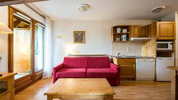 Eine kompakte Küche mit Holzausstattung, einer Mikrowelle und einem Geschirrspüler. Im Wohnbereich befinden sich ein rotes Sofa und ein Holztisch.