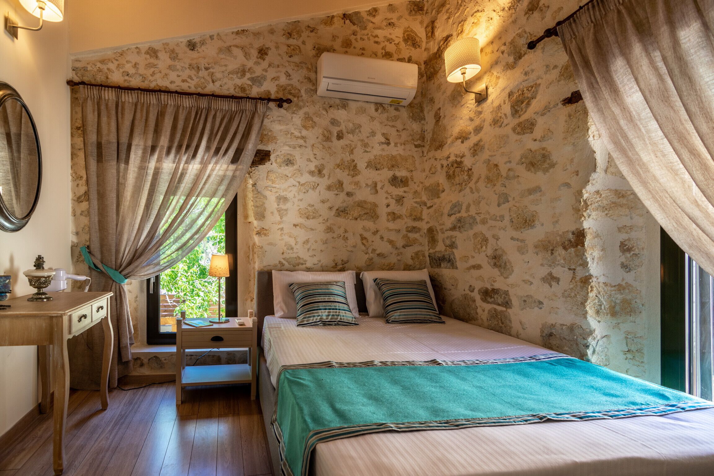 Un dormitorio con pared de piedra, una cama con almohadas a rayas, una mesita de noche con lámpara y una ventana con cortinas.