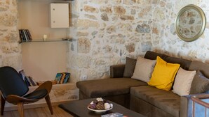 Een gezellige woonkamer met een stenen muur, een bruine bank met geel en beige kussens, een houten salontafel met een schaal snacks en tijdschriften, en een zwarte fauteuil.