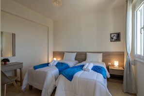 A hotel room with two beds, a desk, a nightstand with a lamp, and a window with curtains. - Haravgi Villa (Rethymno)