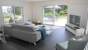 Ein modernes Wohnzimmer mit einer Couch, einem Couchtisch und einem Fernseher auf einem Holzboden. Eine Glastür öffnet sich zu einer Außenpatio mit Stühlen und einem Tisch.