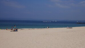 Ein Strand mit Personen, die sich sonnen und spielen, einem klaren Himmel und Segelbooten in der Ferne.