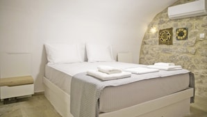 A bedroom with a bed, white bedding, a bedside table, an air conditioning unit, and a stone wall with decorative plates. - Areopolis Art Apartment (East Mani)