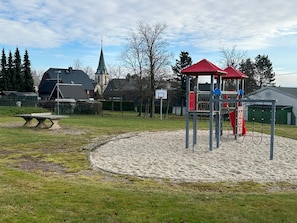 Un terrain de jeux avec une structure de jeu surmontée d’un toit rouge, un panier de basket et une église en arrière-plan.