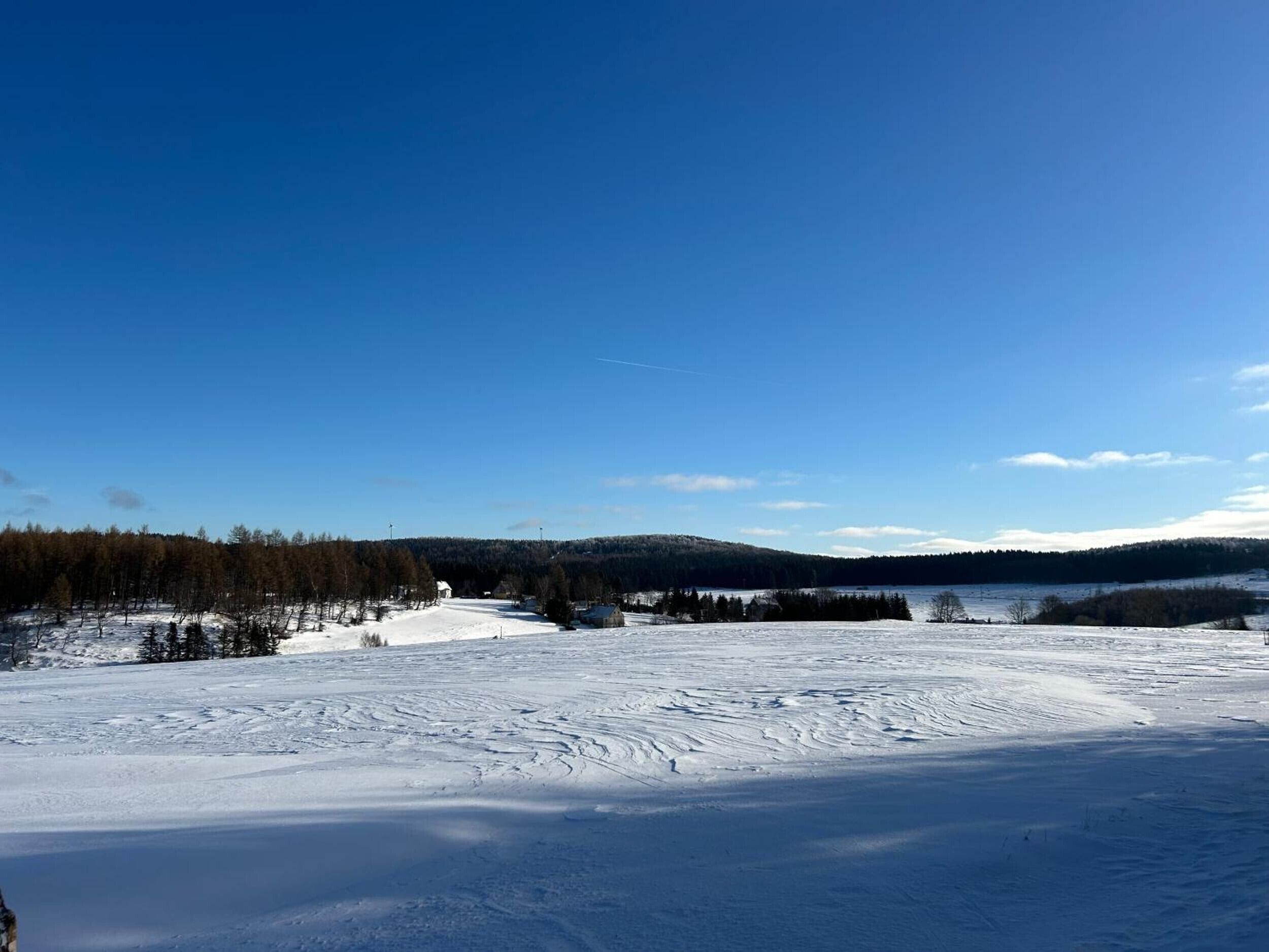 A snowy landscape with a clear sky, a forested hill, and a small building in the distance.