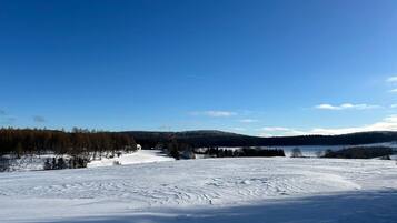 A snowy landscape with a clear sky, a forested hill, and a small building in the distance.