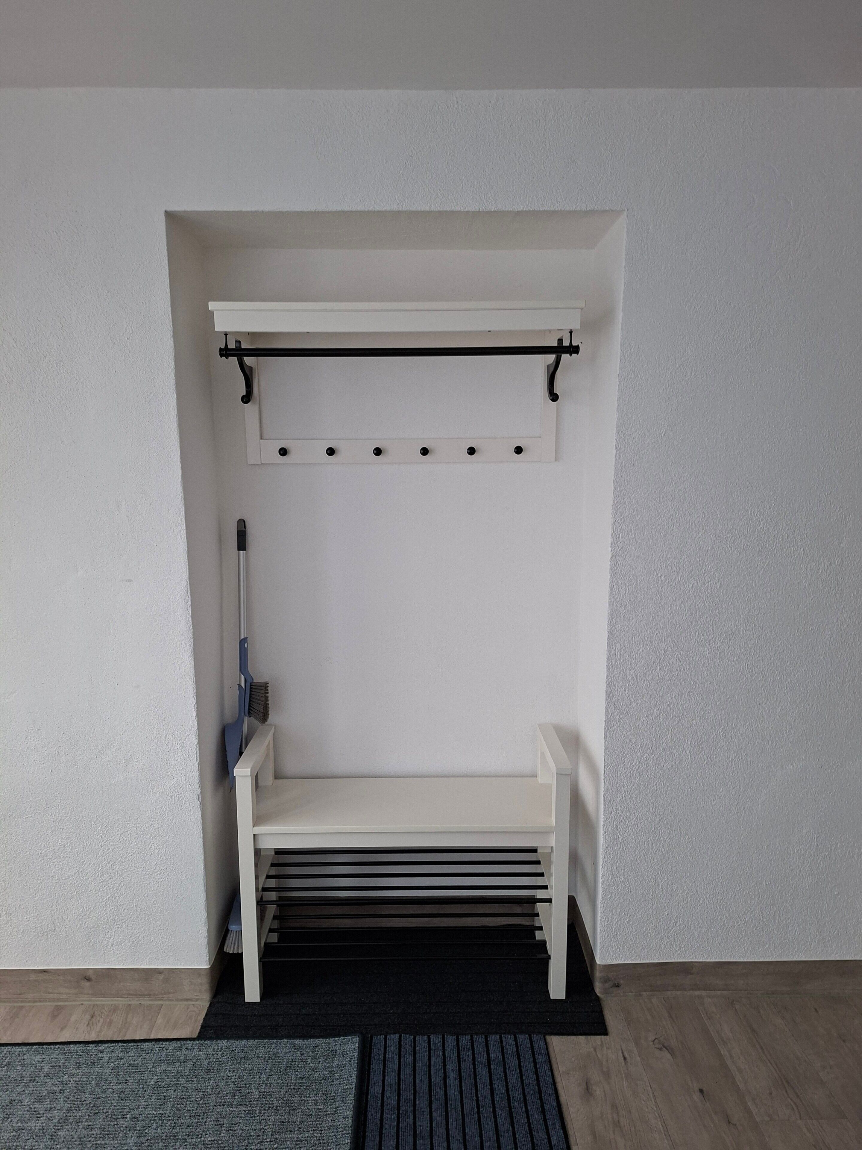 A white bench with a metal rack above it, a broom leaning against the wall, and a black mat on the floor.