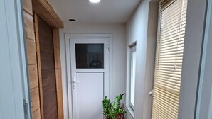 Un pasillo estrecho con suelo de madera, una puerta blanca, una planta en maceta y una ventana con persianas.