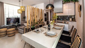 A modern dining area with a long table set for six, surrounded by wicker chairs. A television is mounted on the wall, and there is a kitchen area visible in the background. - Memoria Villa (Rethymno)