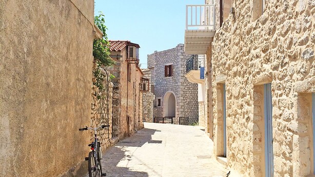 A narrow cobblestone street flanked by stone buildings with balconies, a bicycle parked on the left, and a clear blue sky above. - Areopolis Central Apartment (East Mani)