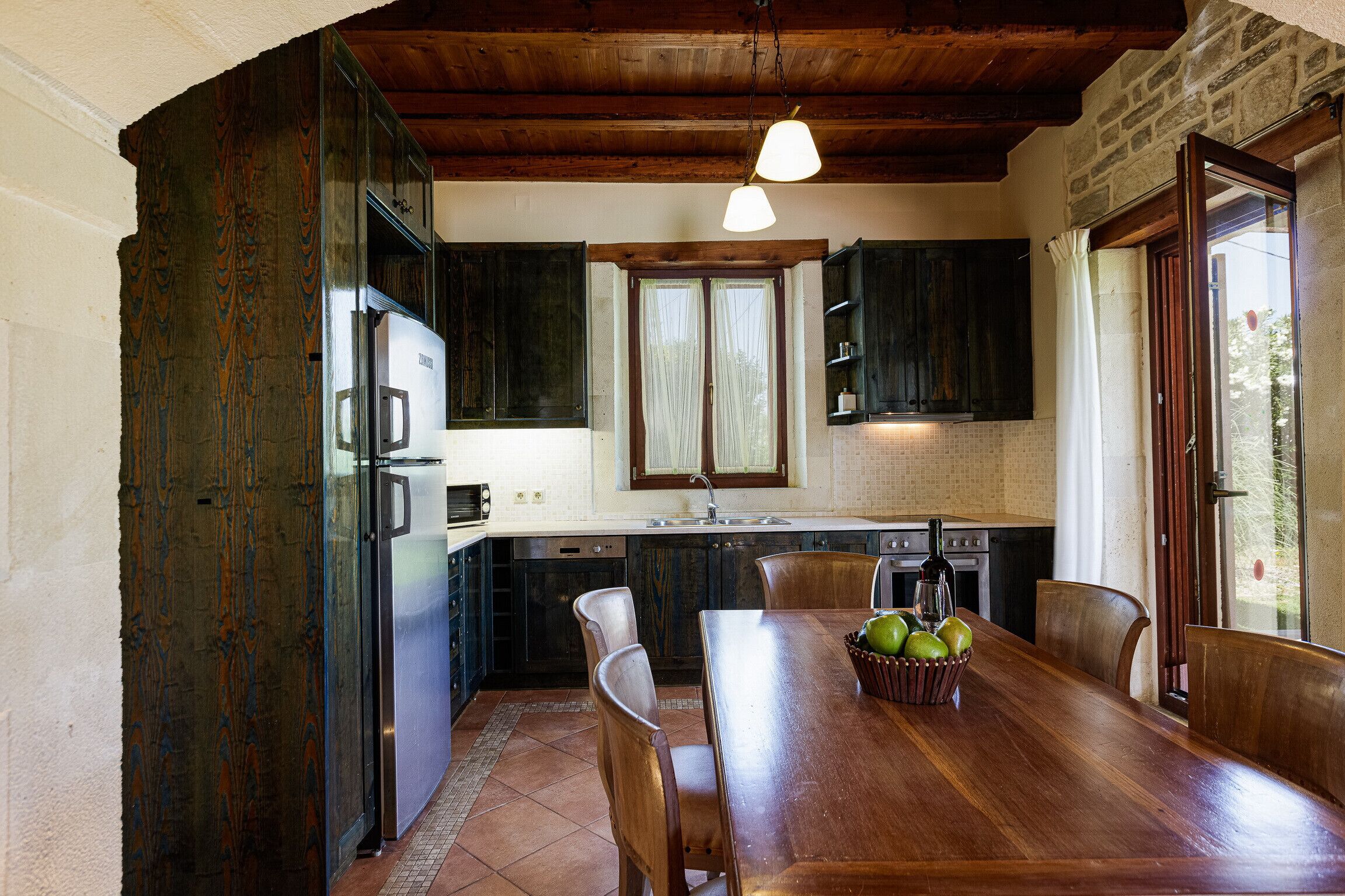 Une cuisine avec des meubles de rangement en bois foncé, un réfrigérateur en acier inoxydable, une table à manger en bois avec des chaises, et une fenêtre avec des rideaux.