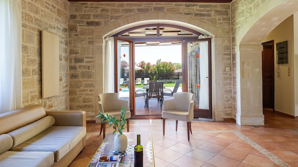 Quarto com paredes de pedra, um sofá, duas cadeiras, uma mesa com um vaso e uma garrafa, e vista através de uma porta arqueada para uma área externa com mesa e cadeiras.
