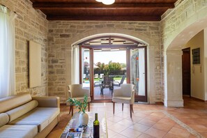 A stone-walled room with a sofa, two chairs, a table with a vase and a bottle, and a view through an arched doorway to an outdoor area with a table and chairs. - Mera Villa (Rethymno)