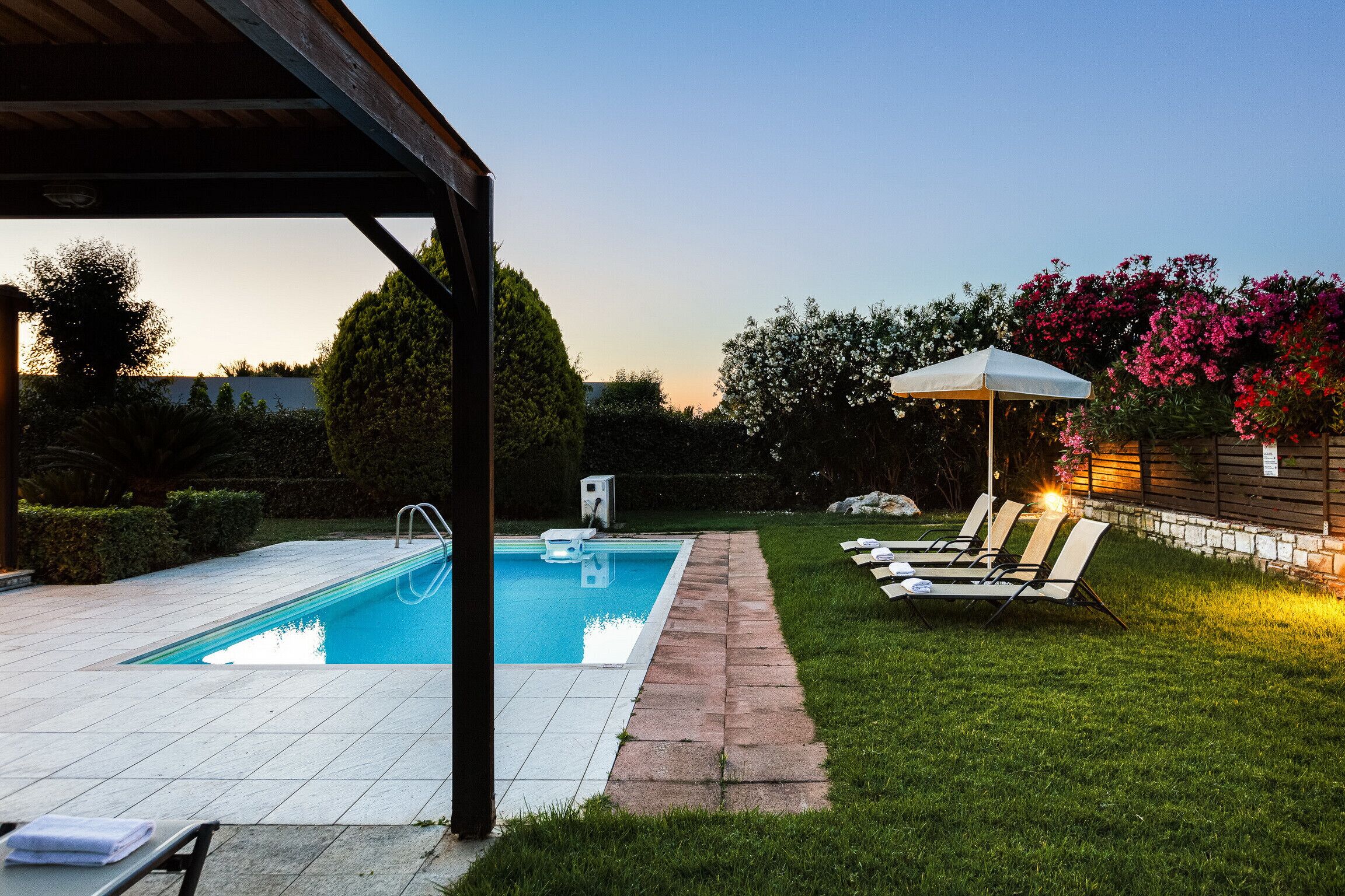 Una piscina con solárium empedrado, un área de descanso techada con sillones de relax y un césped bien cuidado.