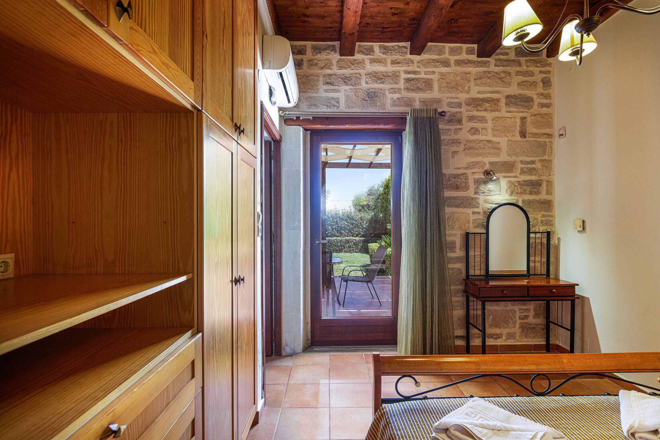 A room with wooden cabinets, a bed with a metal frame, a small table with a mirror, and a view of an outdoor patio with a table and chairs.