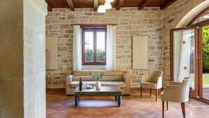 Um quarto com paredes de pedra, teto de madeira, uma janela grande com cortinas, um sofá, uma mesa de centro com um vaso e uma garrafa, e duas cadeiras.