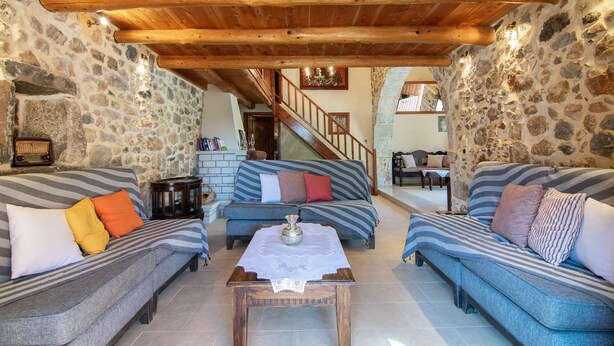 A cozy living room with a stone wall, wooden ceiling beams, and a staircase leading to an upper level. - Archodiko Villa Imbros (Sfakia)