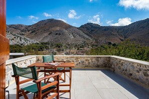 En terrasse med stenvæg, træbord og stole, med udsigt over et bjerglandskab.