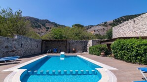 A rectangular swimming pool with a stone wall on one side and a hedge on the other, surrounded by a patio area with lounge chairs. - Archodiko Villa Imbros (Sfakia)