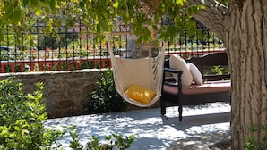 A hammock chair with cushions, a bench, and a tree in a courtyard with a stone wall and metal fence. - Archodiko Villa Imbros (Sfakia)
