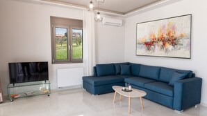 Een moderne woonkamer met een hoekbank, een glazen salontafel, een flatscreen televisie op een glazen voetstuk en een groot abstract schilderij aan de muur.