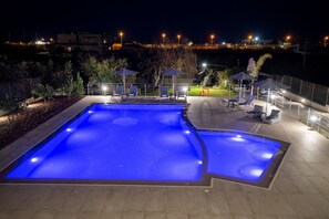 Un área de piscina nocturna con iluminación azul, sillones de descanso y sombrillas.