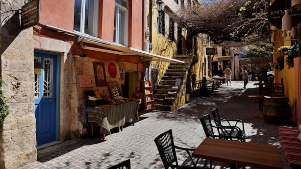 Una calle estrecha flanqueada por edificios, con mesas al aire libre y una escalera que conduce a un piso superior.