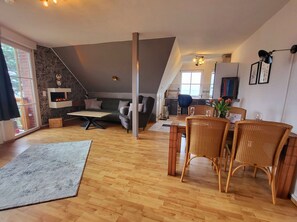 A modern living room with a fireplace, a dining area with wicker chairs, and a kitchen in the background. - Apartment Blick auf Vilm - quietly located apartment with sea view - (Putbus)
