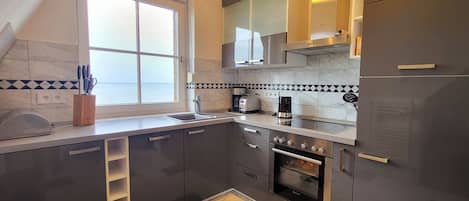 A modern kitchen with dark cabinets, a built-in oven, a coffee maker, and a window overlooking a body of water.