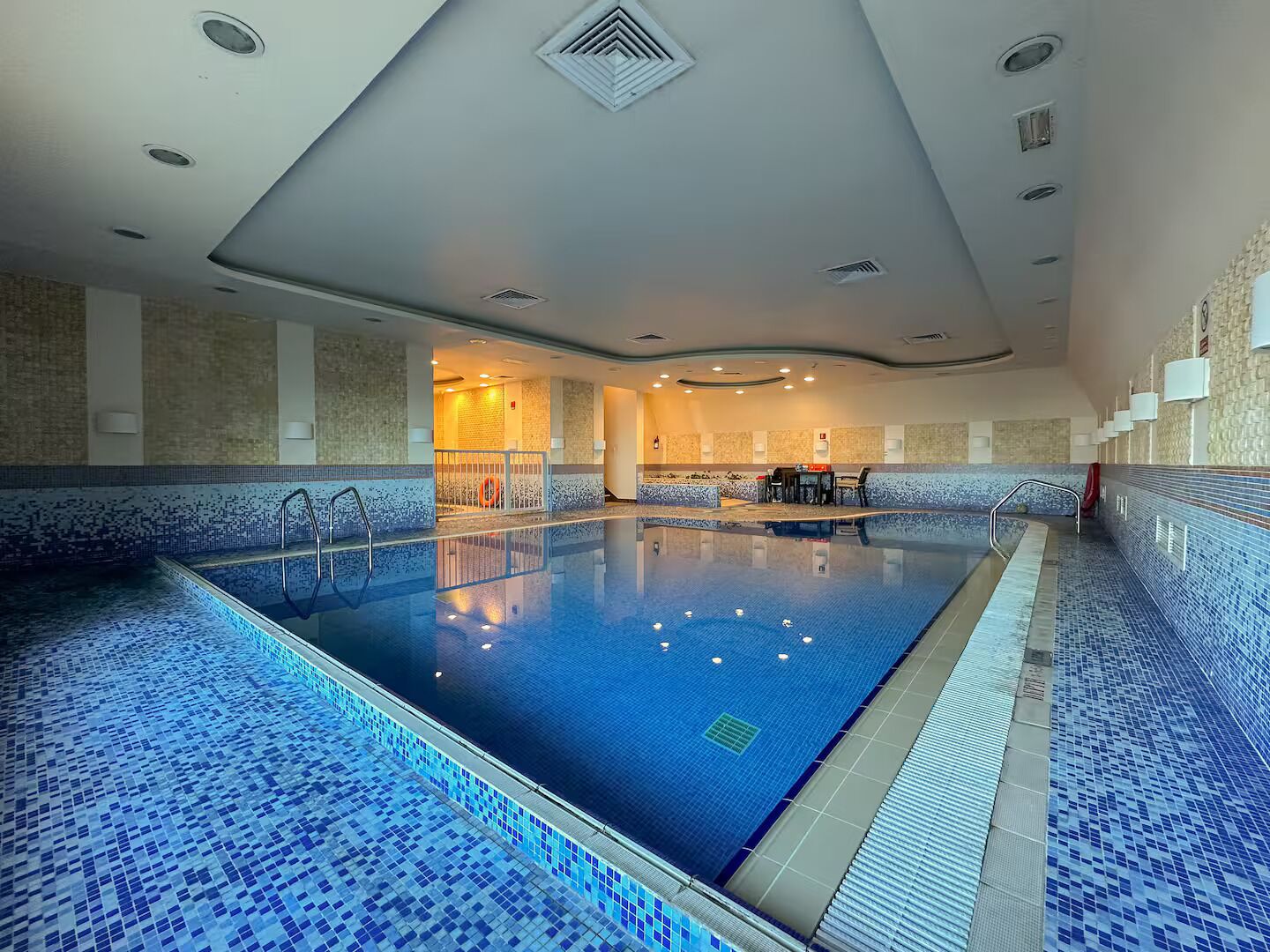 A large indoor swimming pool with blue mosaic tiles, a lifeguard chair, and a seating area with tables and chairs.