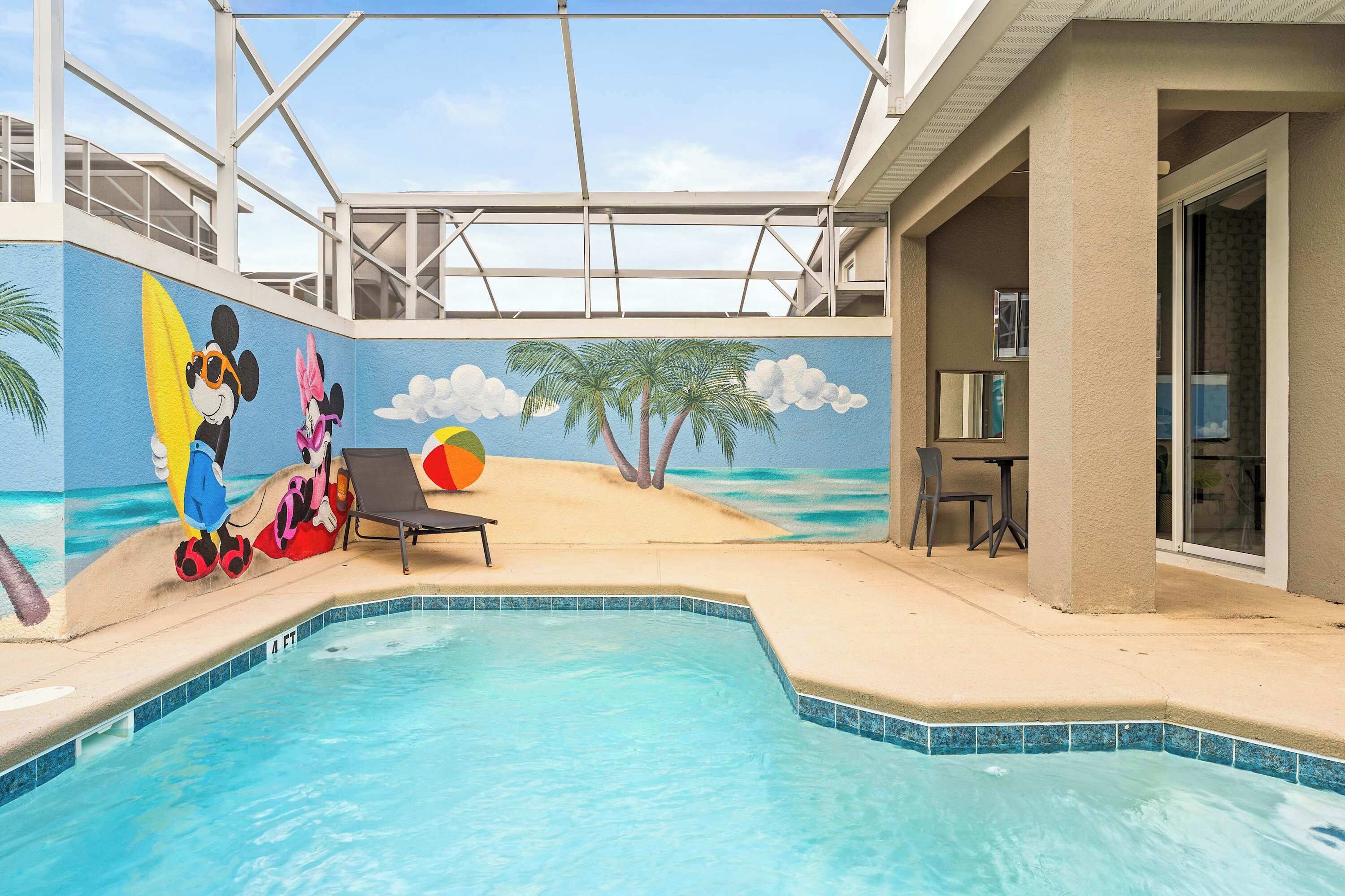 An indoor swimming pool with a mural of Mickey and Minnie Mouse on the wall, a beach scene, and a lounge area with a table and chairs.
