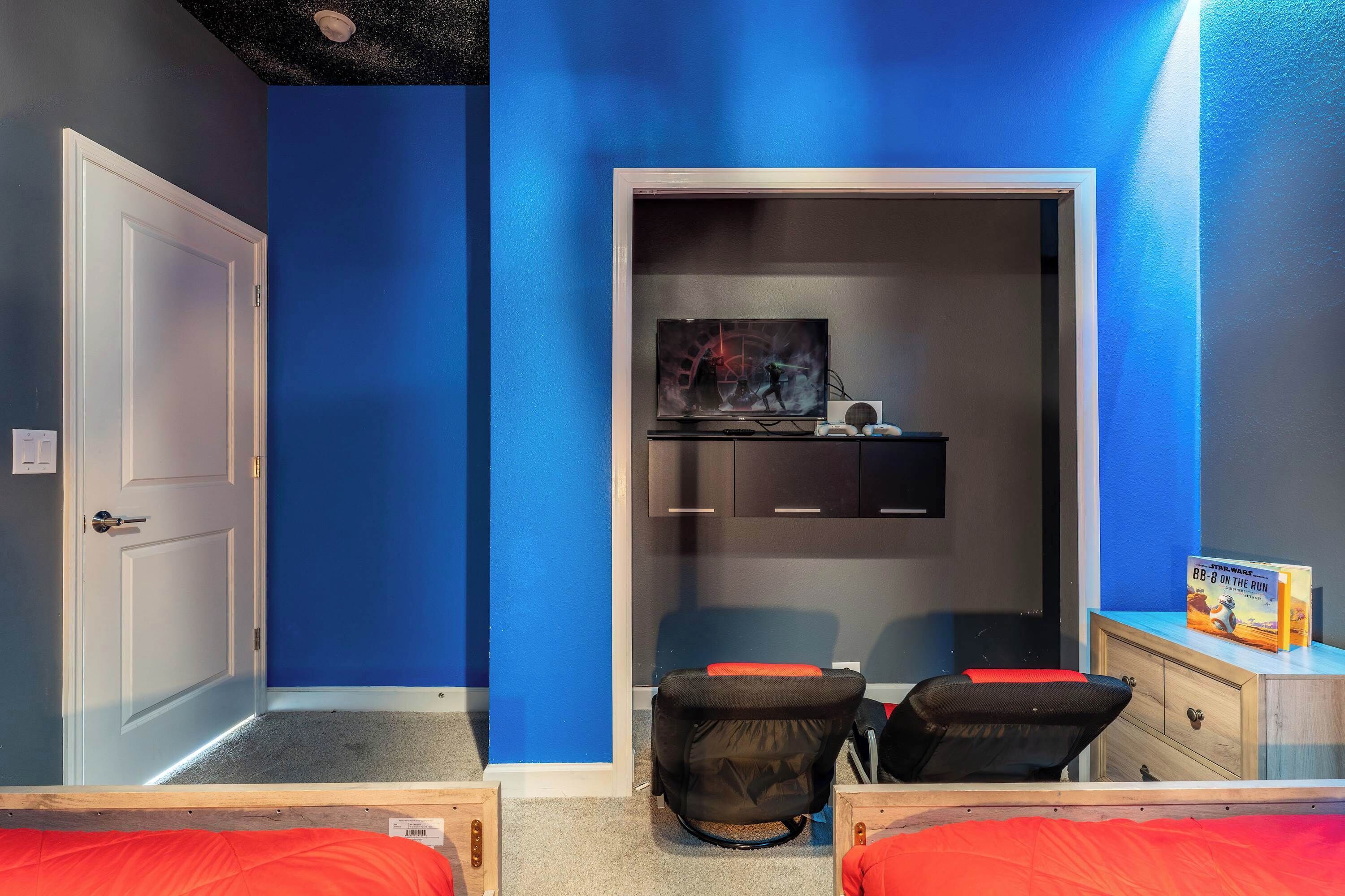 Ein Schlafzimmer mit blauer Wand, einer weißen Tür, einem Bett mit roter Bettwäsche, einem Schreibtisch mit Stuhl und einem Bücherregal.