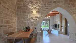 A stone-walled room with a wooden dining table and chairs, a television mounted on the wall, and a view of the outdoors through a large window. - Rosemary Villa (Rethymno)
