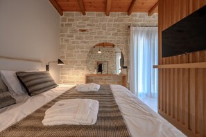 A bedroom with a stone wall, wooden ceiling beams, a bed with white and patterned bedding, a bedside lamp, a flat-screen TV, and a wooden partition. - Rosemary Villa (Rethymno)