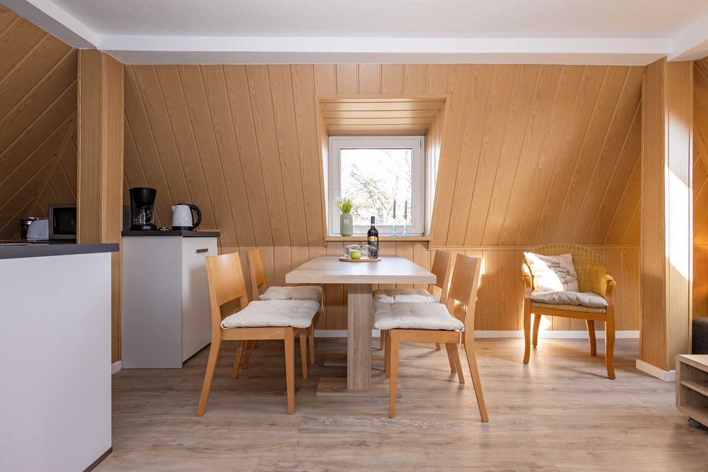 Ein Essbereich mit Holztisch und Stühlen, einem Fenster darüber und einer kleinen Küchenzeile links.