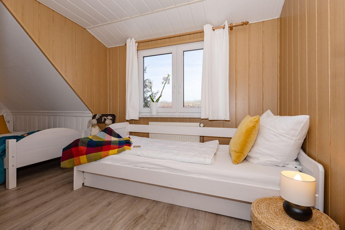Quarto com cama branca, janela com cortinas, luminária e uma pequena planta no parapeito.
