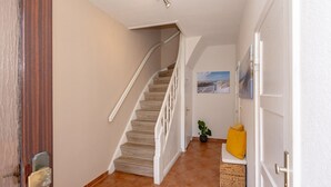 Un pasillo con escalera de madera, una puerta con tirador de latón, una pequeña planta en maceta y un cojín amarillo en una silla de mimbre.