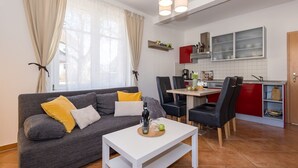 Una sala de estar moderna con un sofá gris, cojines amarillos y una mesa de centro blanca. Se ve una cocina con gabinetes rojos y un comedor con sillas negras al fondo.
