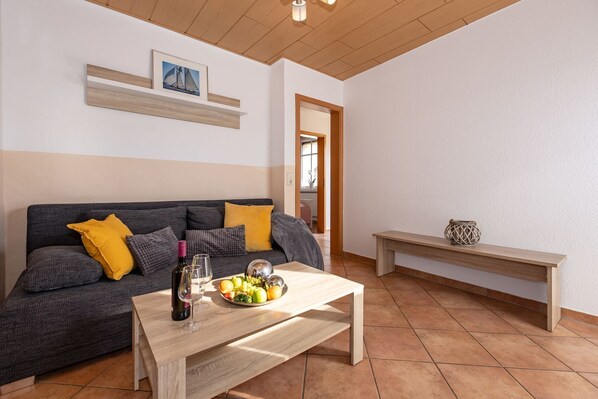 A living room with a sofa, a coffee table with a bottle and glasses, and a fruit bowl. - Harmonie Vacation Rental - Harmonie Vacation Apartment in Rerik - Comfortable (Rerik)