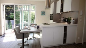 A modern kitchen with white cabinets, a built-in microwave, and a small island. There is a dining area with a table and chairs, and a balcony with a view of greenery. - Linden Palais We 22 - Comfortable stay at the Linden Palais We 22 in (Bad Doberan)