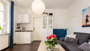Un salon moderne avec un canapé gris, une table basse en bois ornée d’un vase de fleurs, un coin cuisine avec des meubles blancs et un mur d’accent bleu.