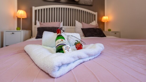 Una cama bien tendida con un edredón rosa, una toalla blanca, una botella de alcohol y una copa con una fresa.
