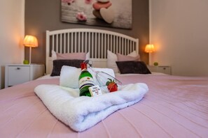 Un lit bien fait, avec une couvre-lit rose, une serviette blanche, une bouteille d’alcool et un verre contenant une fraise.