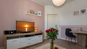 Una sala de estar moderna con televisor, parlantes y un mueble de madera. Hay un jarrón con flores rojas sobre una mesa.