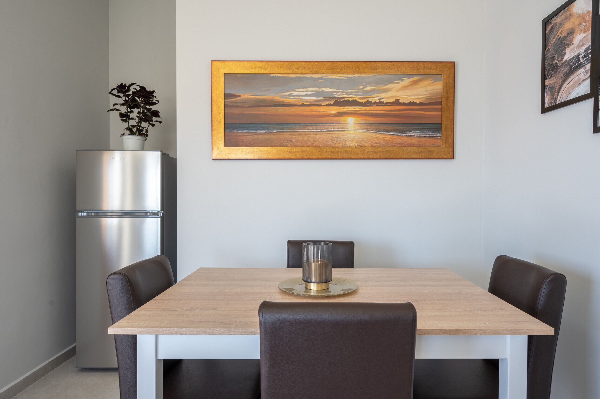 A dining area with a wooden table, four chairs, a stainless steel refrigerator, and a framed painting of a sunset over the ocean.