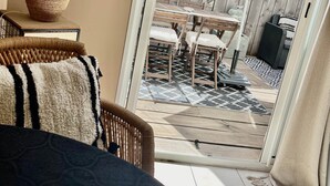 Ein gemütliches Wohnzimmer mit einem Korbstuhl, einem gemusterten Teppich und einem großen Fenster mit Blick auf eine Terrasse, die von einem Holzzaun, Gartenmöbeln und einem dekorativen Sonnenschirm gesäumt ist.