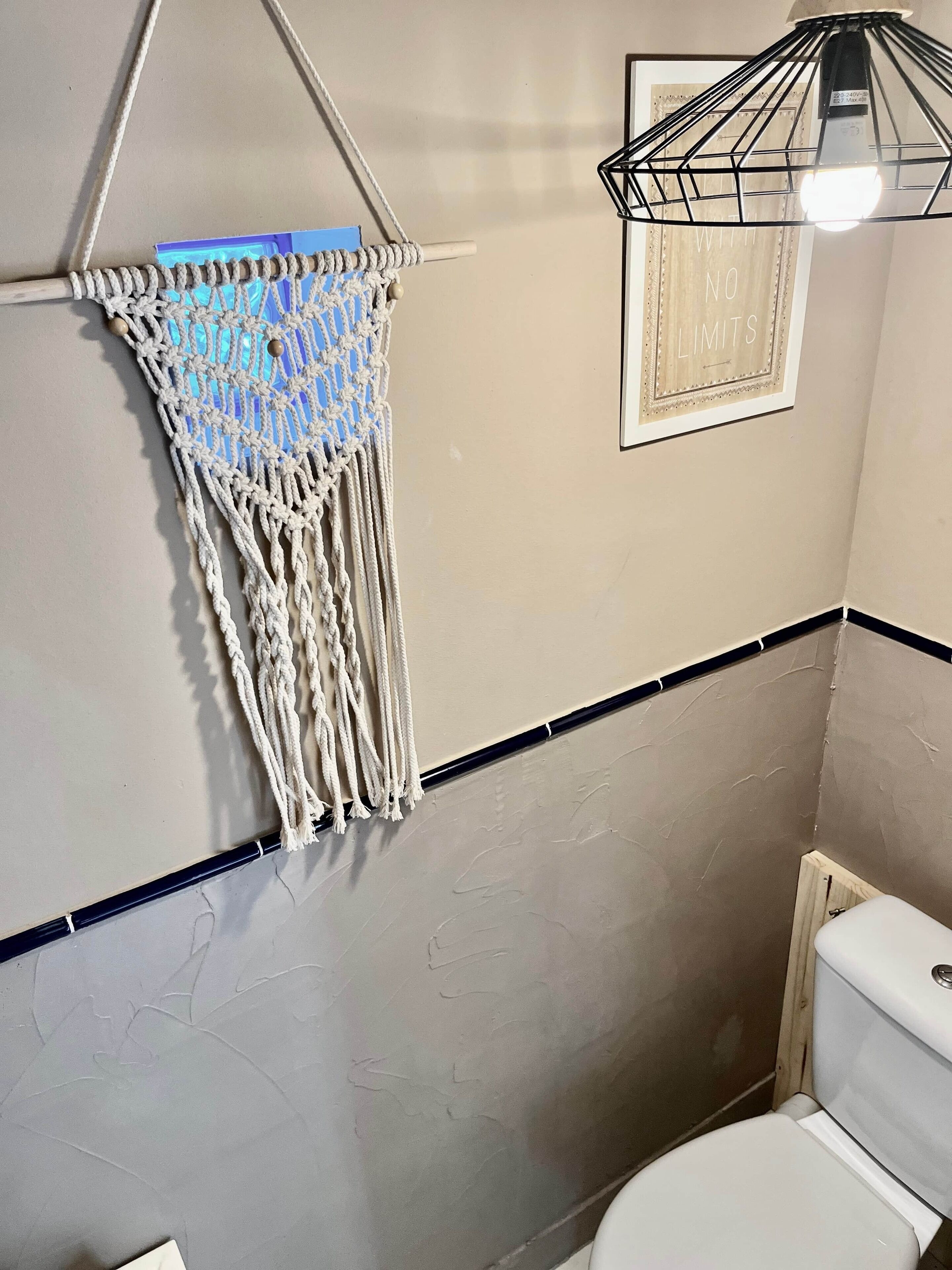 A bathroom with a macrame wall hanging, a framed inspirational quote, and a toilet.