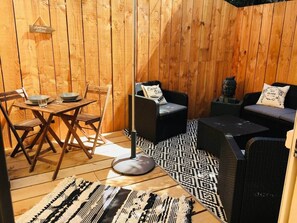 Une terrasse en bois avec une table à manger et des chaises, un lampadaire et deux canapés noirs agrémentés de tapis à motifs.